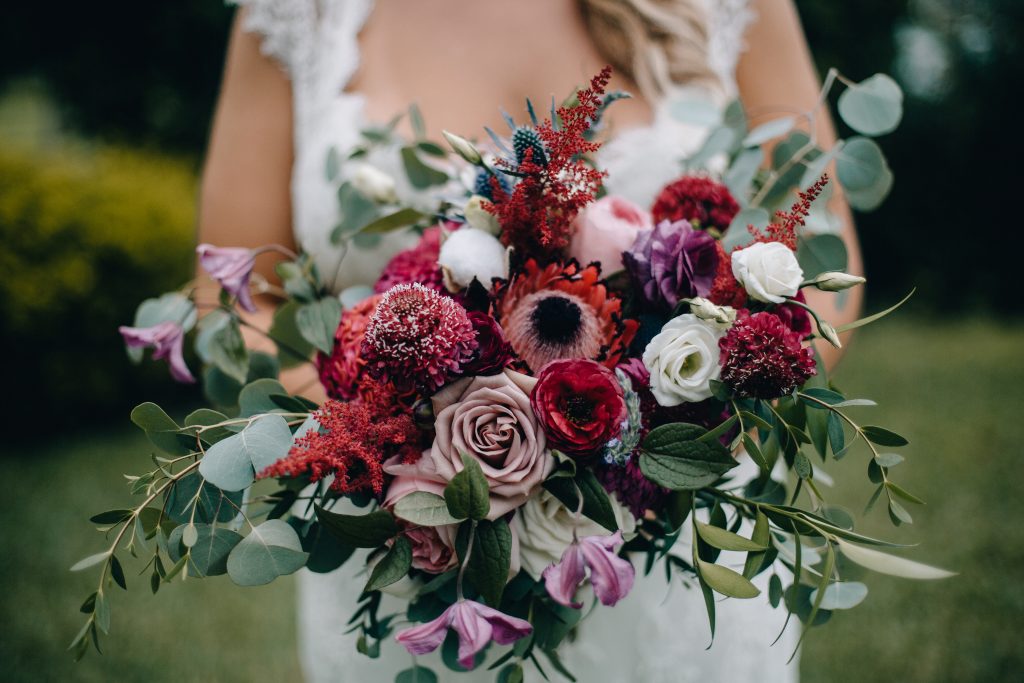 Bohemian textured bridal bouquet in burgundy, purple and raspberry with protea, ranunculus, thistle, lisianthus and roses.