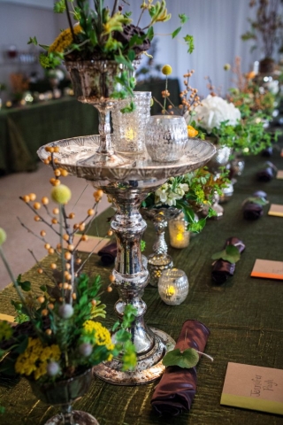 Woodsy centerpieces in silver vases on feast table with candles.