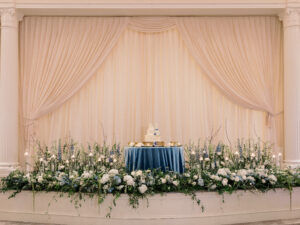 Dramatic garden style stage flowers and cake table of baby blue and white flowers at wedding reception in the Vinoy Grand Ballroom in St. Petersburg, Florida.