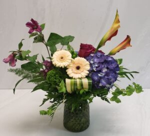 Textured floral arrangement with clematis, calla lilies, and trachelium and more. Arranged in a textured glass vase